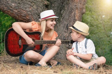 Genç anne ve güzel bir oğlan dinlenme ormandaki şarkı şarkıları bir gitar altında.