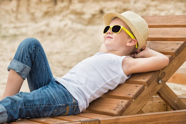 El niño en el sombrero de paja acostado en las tumbonas de madera en la playa.Vacaciones de verano . — Foto de Stock