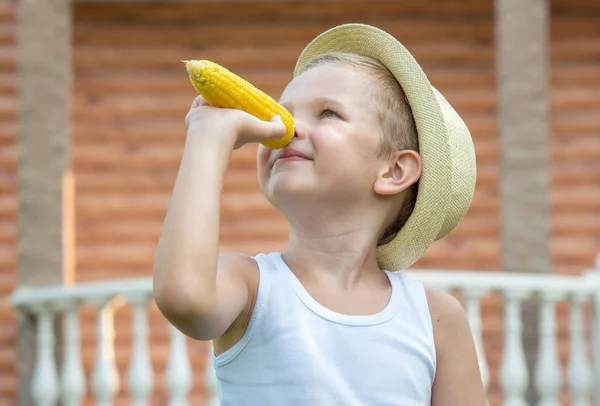 Jongen in stro hoed eten maïs op de kolf in de tuin. Mais stemming. — Stockfoto