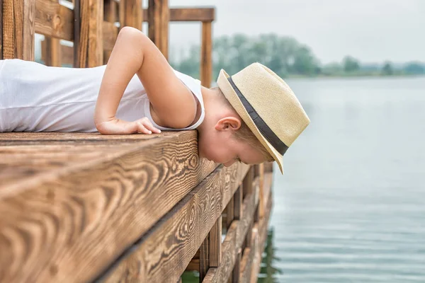 Un niño en un sombrero de paja se encuentra en el muelle de madera y mira el agua. Vacaciones de verano en el lago — Foto de Stock