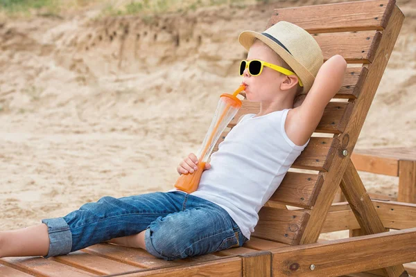 The boy in the straw hat and sunglasses lying on the wooden sun lounger on the beach and drink fresh juice.Summer vacation. — Stock Photo, Image