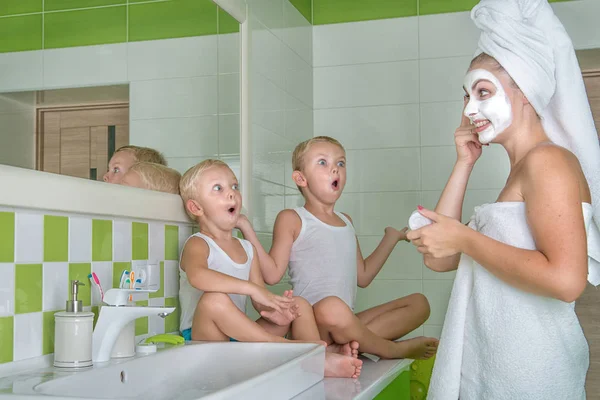 Eine junge Frau machen eine Gesichtsmaske. Kinder beobachten die Mutter, überrascht faces.beauty-Behandlungen für die Haut. — Stockfoto