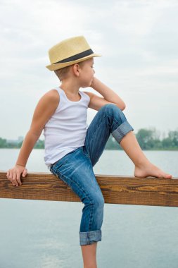 İskelede oturup bir saman şapkalı bir çocuk. Yaz tatili göl kenarında