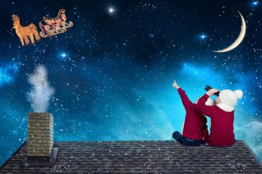 Mutlu Noeller ve mutlu tatiller! Çatıda oturan ve Noel Baba onun kızak ay gökyüzü uçan bakarak iki küçük kardeş. Çocuklar tatil keyfini çıkarın. Noel gösterge kavramı.