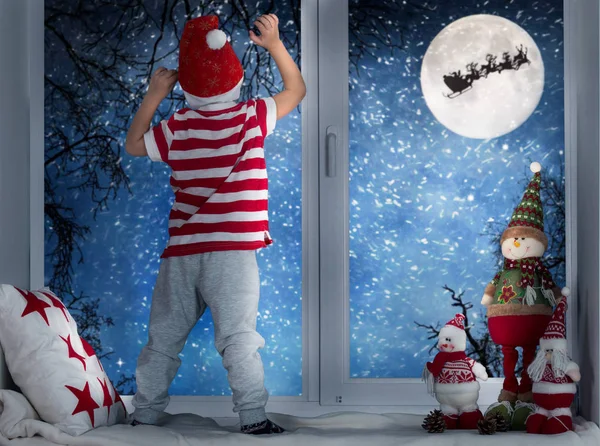 Kellemes karácsonyi ünnepeket és boldog ünnepek! Kis gyermek fiú áll az ablak, és keresi a Mikulás repül a szán Hold ég ellen. Szoba díszített, a karácsony. Gyerek élvezni az ünnep. — Stock Fotó