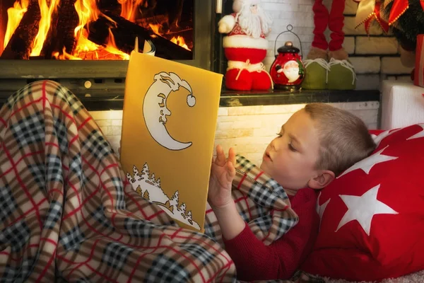 Boldog karácsonyt! Aranyos fiú olvasó egy könyv feküdt a szőnyegen, a kandalló mellett. — Stock Fotó