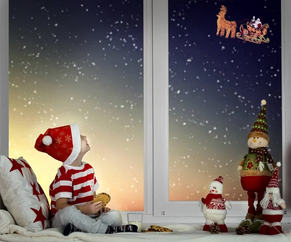 Mutlu Noeller ve mutlu tatiller! Pencerenin üzerinde oturan ve Noel Baba onun kızak ay gökyüzü uçan bakarak küçük çocuk. Noel'de dekore edilmiş Oda.