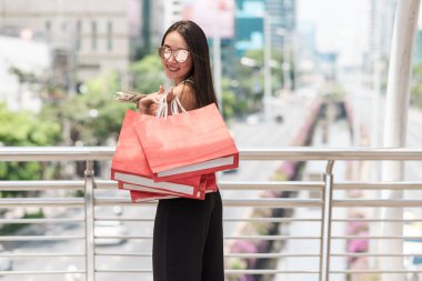 Kentsel şehir kavramı shopper modern tek kadın. Kırmızı alışveriş kağıt torba ve nakit mutlu büyük şehirde alışveriş duygu dolar tutan gülümseyerek Modern yeni zengin genç Asya kadın. Arka planda Road alışveriş ile.