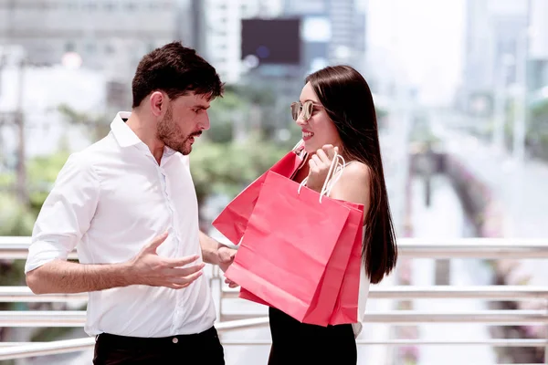 Couple argue after a hard day of huge sale shopping in asian country. Unhappy young handsome white male and beautiful asian girl with shopping bag looking very satisfy as she spent all his money.