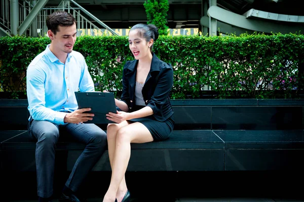 Young business leaders concept. Young Business man discussing work with woman co worker outdoor in city. Smart casual man with blue shirt and smartly dress woman with notepad sitting down talking.