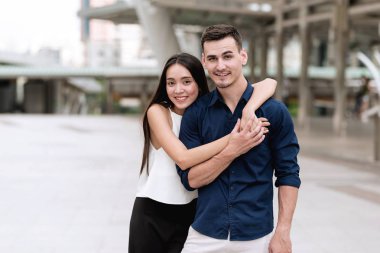 Mutlu genç ırklararası çift. Mutlu çekici genç Asyalı kadın sarılmak yakışıklı bir beyaz adam çok sıkı bir gülümseme ile aşkını onun geleceği için gösterilen açık yerde. Kentsel görünüyor uygulamak.