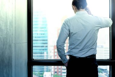 Genç iş lideri kavramı. Genç yakışıklı iş adamı pencereden dışarı bakarak telefon modern ofis. Mobil aygıtta konuşma ile çizgili gömlek beyaz erkek.