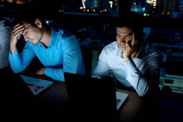 Working overtime concept. Group of young business team working late at night in meeting room trying to complete their assignments. Taken in modern office with office building in background.