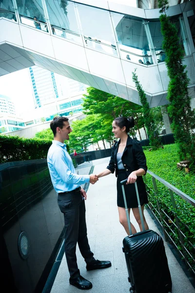 Young business leader concept. Young business man and woman shaking hand in urban city with woman holding a roller bag, vintage look. Smart casual man with blue shirt and smartly dress woman talking.