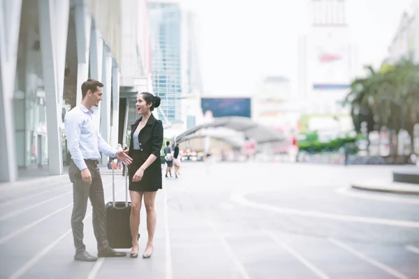 Young business leader concept. Young business man and woman walking down the street in urban city with woman holding a roller bag, vintage look. Man with blue shirt and smart dress woman shaking hand.