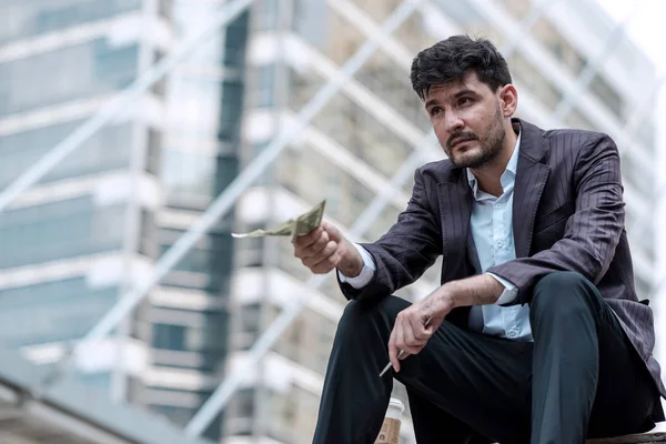 Low income white collar concept.  Handsome white direct sale man sitting down with his hard earn dollar cash after a hard day of direct selling. With sky walk and office building in background.