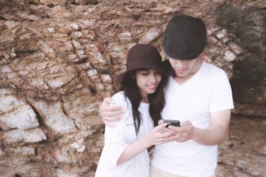 Hareket eden telefon kayalık plaj arka plan ile oynarken bir çift. Genç mutlu ırklararası çift sahilde cep telefonu arıyorum. Asyalı kadın, beyaz adam. Genç karışık yarış romantizm kavramı.