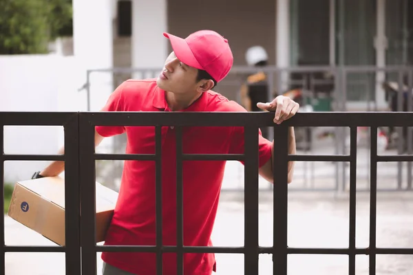 Young logistic career concept. Happy delivery man looking for his customers at their home. Taken in real house. Asian chinese fit man in red polo shirt and jeans with red hat in his early twenties.