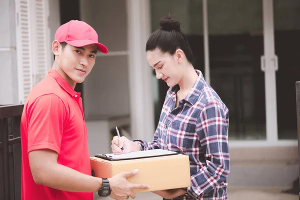Young logistic career concept. Happy delivery man giving his package to customer at home. Taken in real house. Asian chinese fit man in red polo shirt and jeans with red hat in his early twenties.