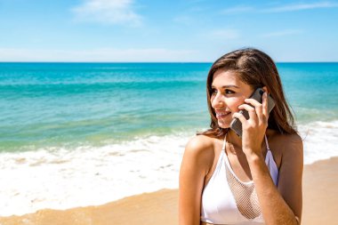 Güzel genç tek beyaz kadın plajda. Cep telefonu üzerinde beyaz bikini giyen, konuşurken. Bağlı dünya kavramı.