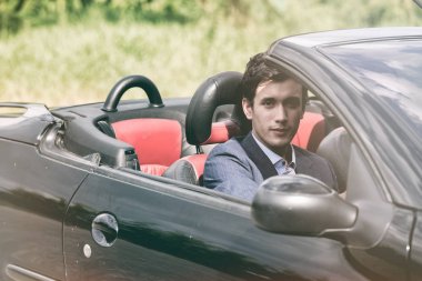 Tatil araba kira ve yol yolculuk kavramı. Kamera, bir yolculuğa ve uzun yolculuk için hazır bakarak yakışıklı beyaz adam doğa arka plana sahip siyah bir Cabrio içinde.