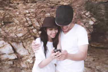 Hareket eden telefon kayalık plaj arka plan ile oynarken bir çift. Genç mutlu ırklararası çift sahilde cep telefonu arıyorum. Asyalı kadın, beyaz adam. Genç karışık yarış romantizm kavramı.