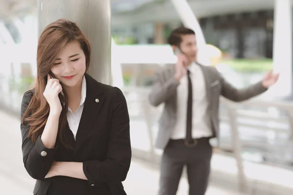 Confident young business woman in smart office dress on phone. Beautiful white asian female worker in black jacket with brown hair talking on phone with asian man in grey suit in background.