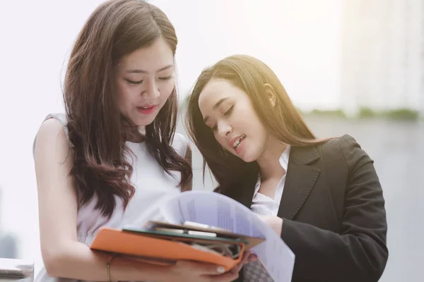 Two beautiful confident asian business intern woman taking to each other. Two women in office suit talking during office break time, taken outdoor in natural light. Career woman concept.