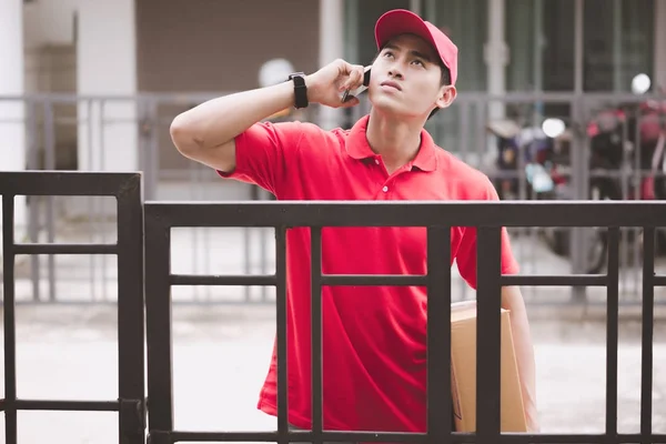 Young logistic career concept. Happy delivery man in front of customer house calling them to get the package. Taken in real house. Asian chinese fit man in red polo shirt in his early twenties.