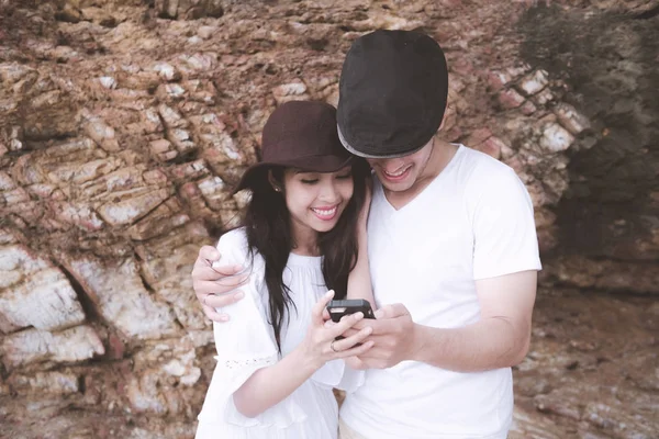 Hareket eden telefon kayalık plaj arka plan ile oynarken bir çift. Genç mutlu ırklararası çift sahilde cep telefonu arıyorum. Asyalı kadın, beyaz adam. Genç karışık yarış romantizm kavramı.