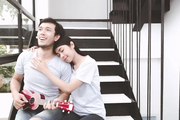 Mixed race lovers concept. Young white male playing banjo to his pretty young chinese girlfriend on the stairs. Wearing blue jeans, blue shirt and in their early twenties.