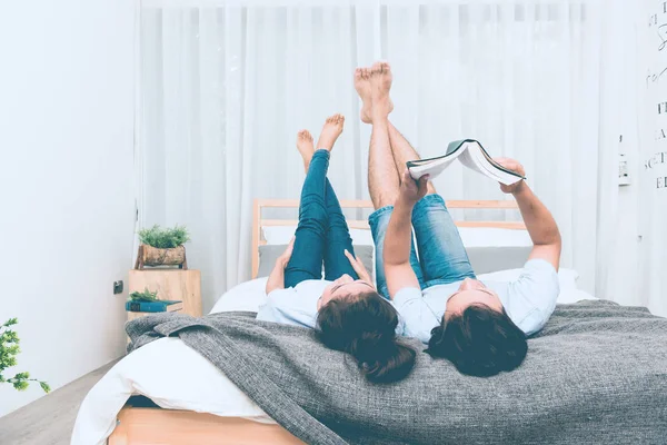 Mixed race lovers concept. Young white male reading a book in his bedroom with his pretty young chinese girlfriend to join. Wearing blue jeans, blue shirt and in their early twenties. Taken indoor.