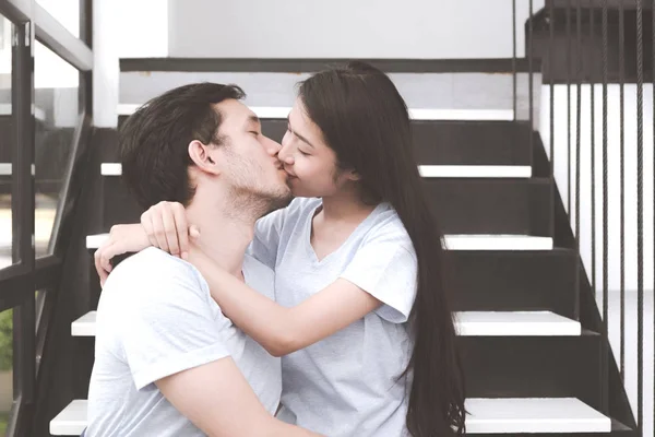 Mixed race lovers concept. Young white male kissing his pretty young chinese girlfriend on a stairs. Wearing blue jeans, blue shirt and in their early twenties. Taken indoor.