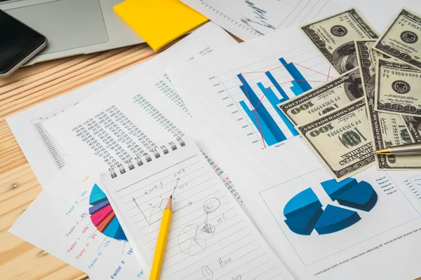 Фінансові графіки на столі з ноутбуком, телефоном, доларовим банком — стокове фото