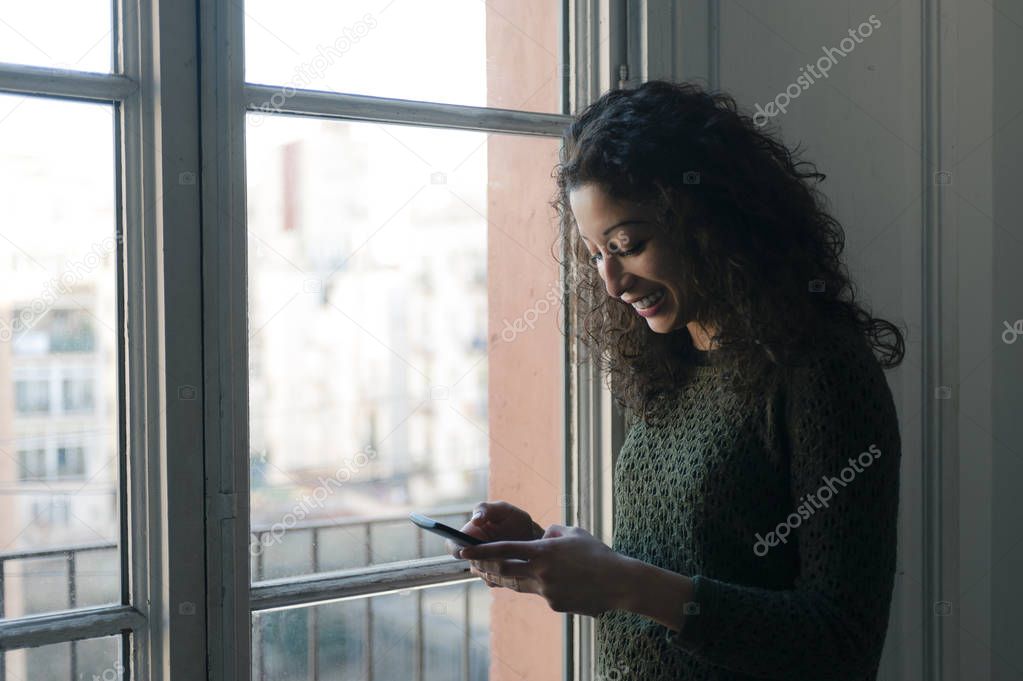 Latin woman chating with her mobile phone next to a window at home