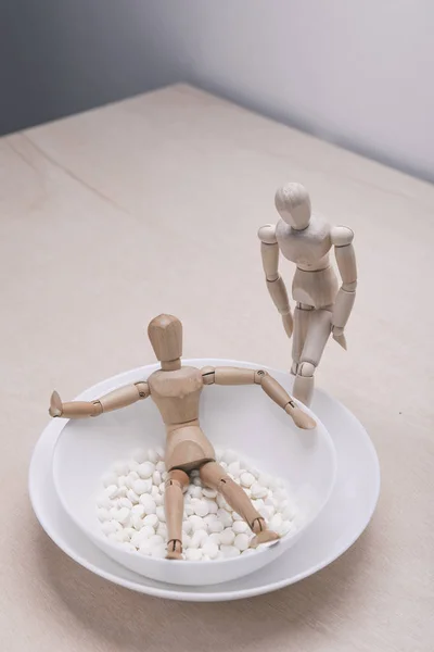 wooden mannequin lying in bowl with pills, another mannequin standing near bowl