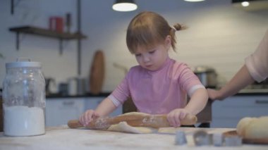 Mom teaches daughter to cook the dough. The girl learns to cook pastries. Kneading the dough together. Roll out the dough for baking. A wonderful family in the spacious kitchen prepares food. Holiday cooking sweets. Little helper in the kitchen.