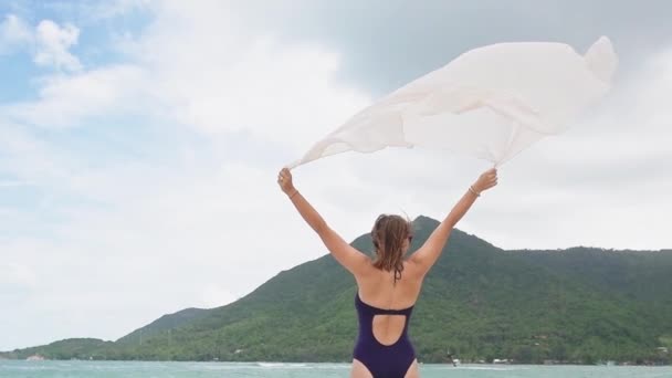 海滨有一条围巾的女孩 海风风吹在头发上 在热带海滩上穿着蓝色泳衣的自由女人 一条白色的丝巾在风中飘扬 — 图库视频影像