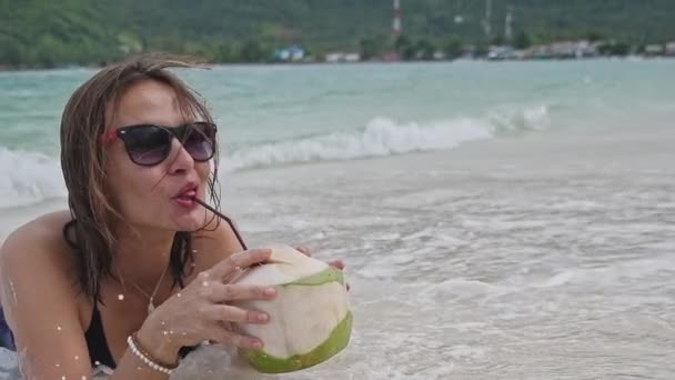 一个年轻的女人躺在沙滩上 女孩在喝椰子水 椰子酱鸡尾酒 在热带岛屿上微笑的女人浪花中的女孩 — 图库视频影像