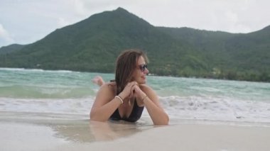 Kumsalda mayo giymiş genç ve güzel bir kız kumsalda yatıyor. Ayaklar havada. Tropik bir adada kumsal. Beyaz kumlu sahilde oynaşan bir kız.