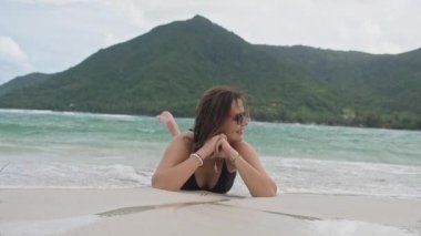 Kumsalda mayo giymiş genç ve güzel bir kız kumsalda yatıyor. Ayaklar havada. Tropik bir adada kumsal. Beyaz kumlu sahilde oynaşan bir kız.