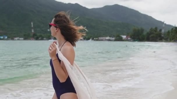 一个女孩在热带海滩上手牵着一个长袍 一位身穿泳衣 戴着丝巾的年轻女子在海滨散步 — 图库视频影像