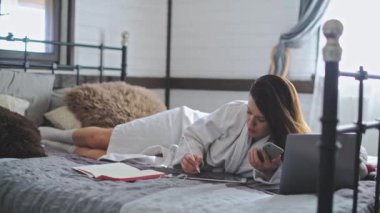 Genç, güzel bir kız bornozla yatakta yatıyor. Dizüstü bilgisayarda çalışan güzel bir kadın. Evde güneşli bir sabah. Elektronik cihazlar. Çizim için tablet.