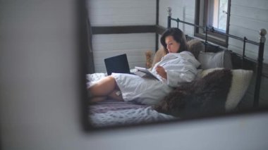 Genç, güzel bir kız bornozla yatakta yatıyor. Dizüstü bilgisayarda çalışan güzel bir kadın. Evde güneşli bir sabah. Elektronik cihazlar. Çizim için tablet.