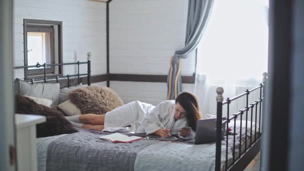 若く美しい少女がベッドの上のバスローブに横たわっている ラップトップで働くかなりの女性 家での晴れた朝 電子機器だ 図面用タブレット — ストック動画