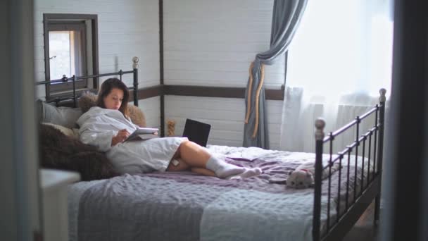 若く美しい少女がベッドの上のバスローブに横たわっている ラップトップで働くかなりの女性 家での晴れた朝 電子機器だ 図面用タブレット — ストック動画