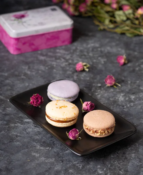 Koyu arkaplanda pembe ve mor badem kurabiyeleri ve gül çiçekleri. Makaronlar yaklaşsın..