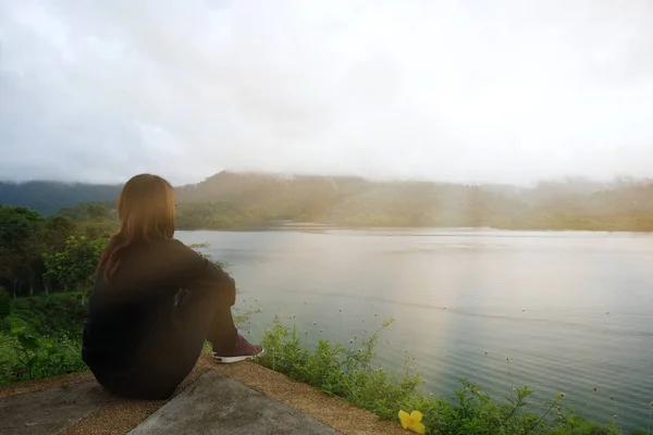 Mujer sentada sola en el acantilado. frente a ella tienen mar y la hora de la puesta del sol son el fondo. esta imagen para el paisaje, vacaciones y concepto de la naturaleza —  Fotos de Stock