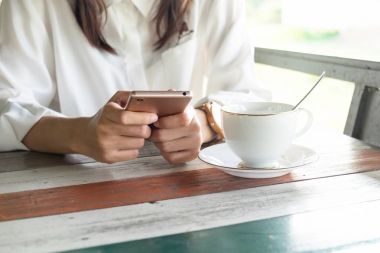 Önden Görünüm. cep telefonu almak sırasında bir ara sabah oynayan genç kadın. ön kahve fincanı ahşap masanın üzerine koyarak sahip. Bu görüntü için iş, teknoloji, içecek ve portre kavramı