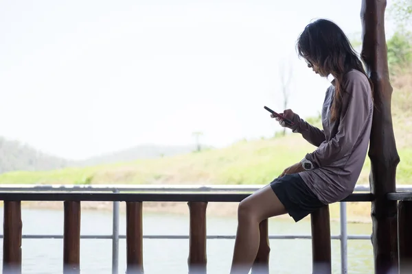 Mujer solitaria jugando al teléfono móvil mientras está sentado balcón de madera en el paseo marítimo. esta imagen para viajes, retrato y tecnología —  Fotos de Stock